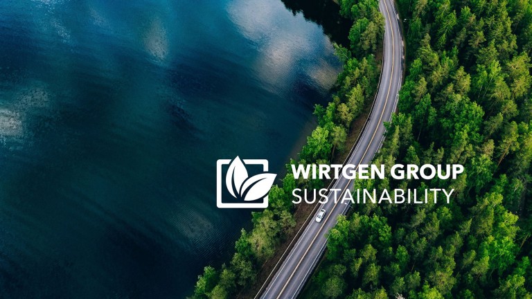 The tagline ‘Wirtgen Group Sustainability’ in white superimposed on a view of a car driving through green woods on a lakeside road