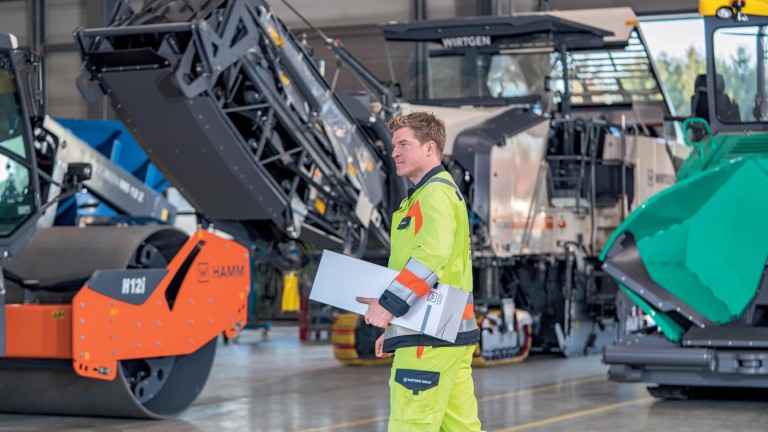 Service technician with a spare part in a workshop with construction equipment standing in the background