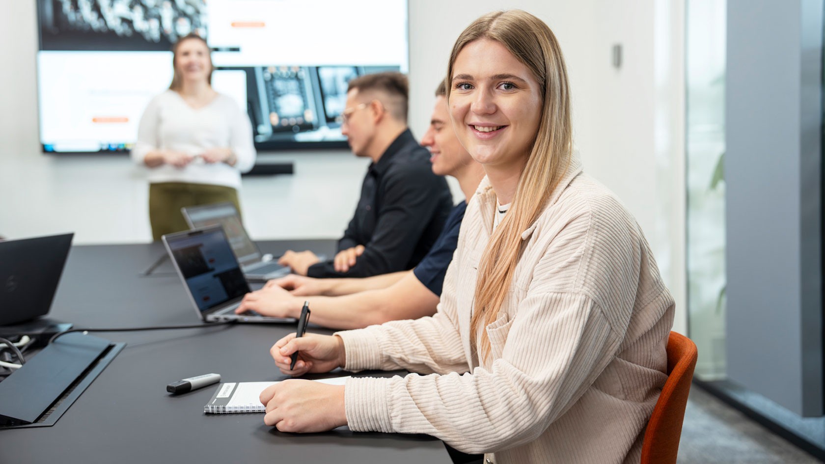 Lachende Studentin sitzt mit Schreibmaterial an einem Tisch mit mehreren Studenten