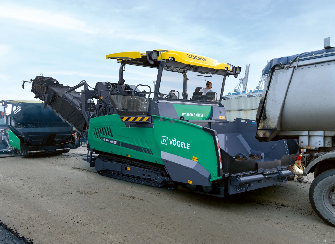 The photo shows a VÖGELE PowerFeeder transferring material to a road paver on a construction site.