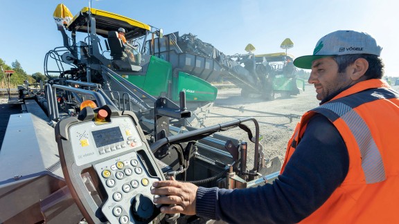 Paver operator operating ErgoPlus 3 screed console in the foreground, two Vögele pavers can be seen in operation in the background