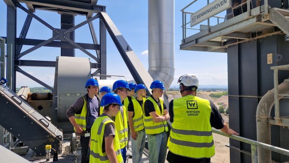 9 Auszubildende mit blauem Helm und ein Ausbilder mit weißem Helm stehen mit gelber Warnweste auf einer Platztform hoch oben auf einer Benninghoven Mischanlage