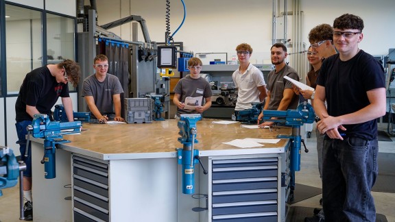 Acht junge Auszubildende stehen rund um eine achteckige Werkbank in der Werkshalle bei Benninghoven