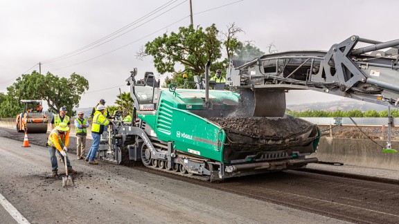Vögele Super 2000-3i being loaded with milled material by the milling machine and Hamm roller compacting in the background