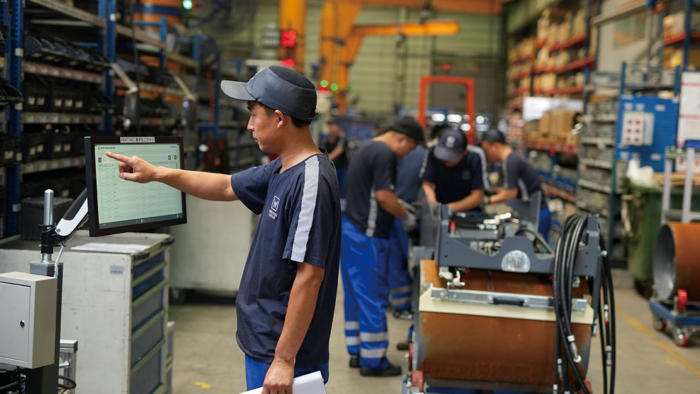 Chinese employee in production taps a display in the foreground with his right index finger, while his colleagues in the background continue to work on a Hamm roller