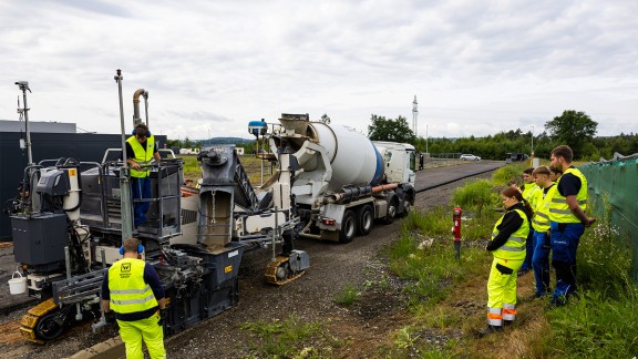The trainees stand on a rise beside a road and watch a Wirtgen slipform paver at work.