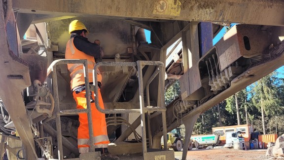 Находясь на рабочей площадке дробилки, оператор выполняет замену сита на первичном грохоте. По периметру площадки установлен поручень.