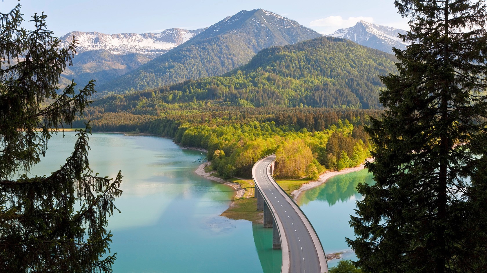 Una carretera atraviesa un lago en un bosque idílico con montañas en el fondo.