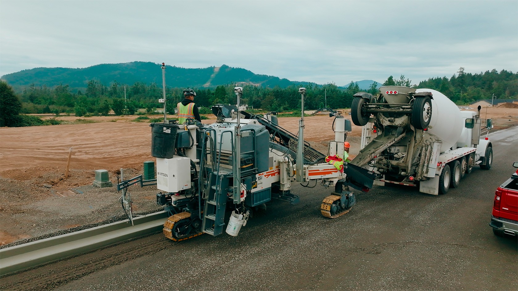 Vue légèrement latérale sur la SP 15i sur une route départementale lors de la pose de bordures de trottoir. À l’arrière-plan, des champs et des collines boisées.