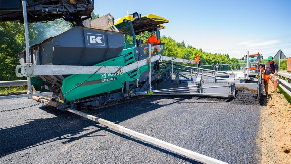 The paver from the front, the operator’s seat has been swung out and extends out over the road surface for a better overview.