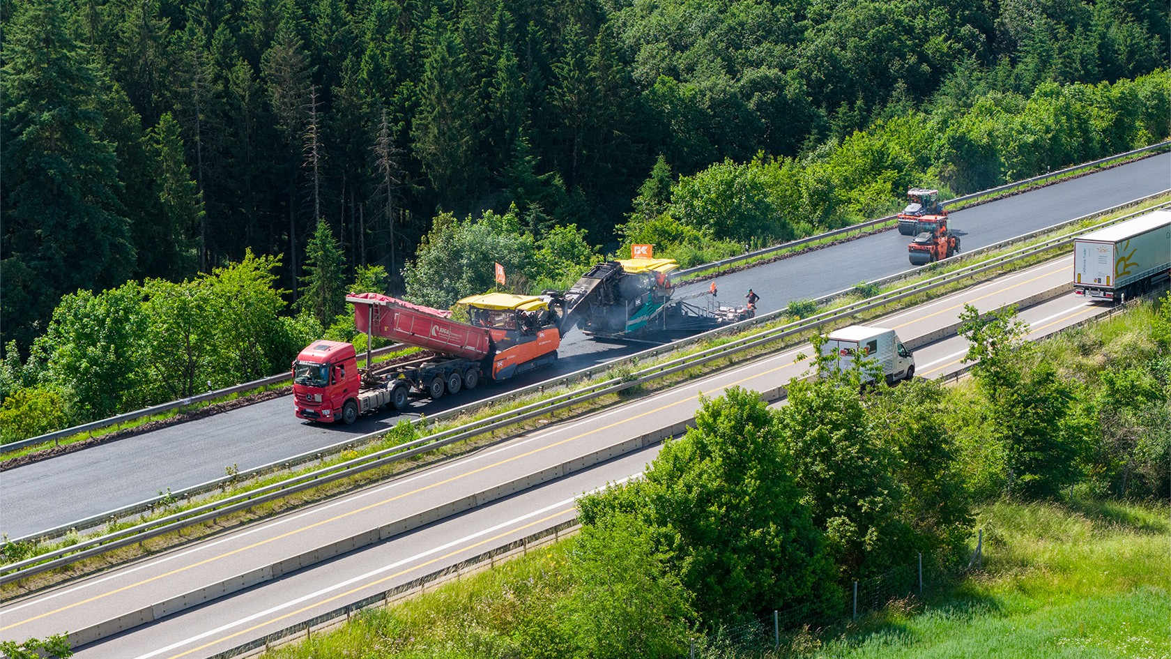 Aerial photograph of a paving train consisting of a mobile feeder, a paver and rollers rehabilitating one lane of a highway embedded in a green landscape.