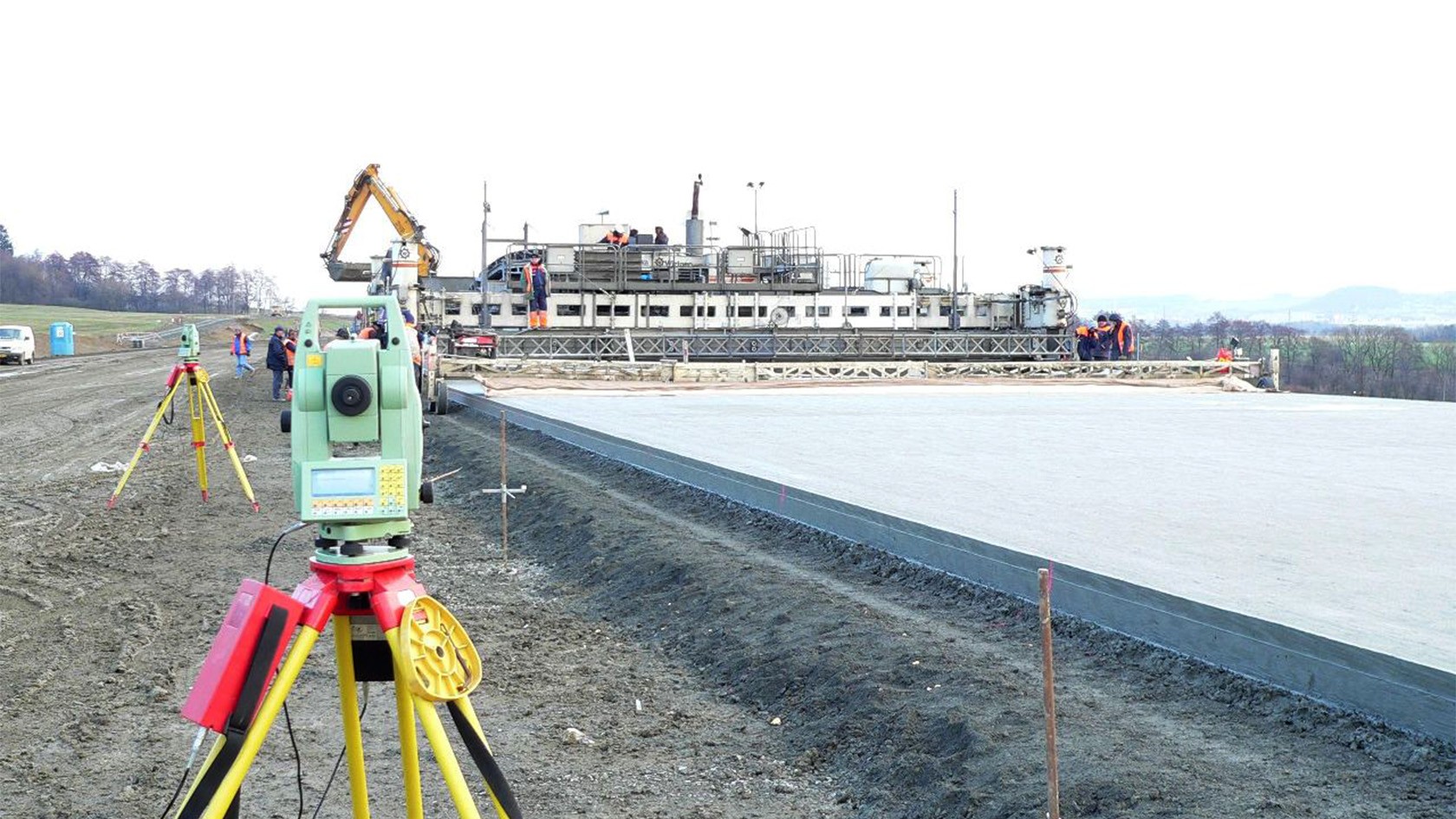 A concrete paving project with a total station in the foreground and an inset slipform paver in the background.