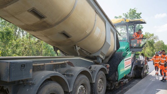 A tipper truck unloads recycling mix to a Vögele paver for paving.