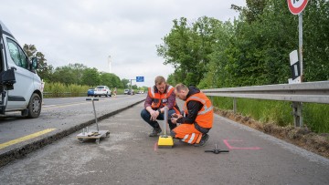 Two men in hi-vis safety vests crouch on the first compacted paved layer with technical instruments