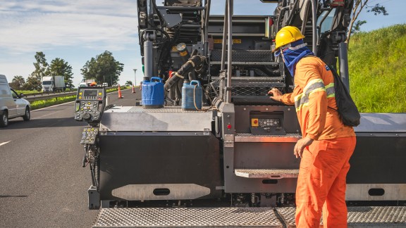 Worker stands behind the SUPER 2100-5 X and operates Paver Access Control (PAC) from the screed control unit.