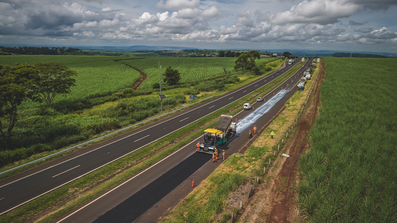 Bird’s-eye view of a two-lane motorway surrounded by fields of sugar cane. The SUPER 2100-5 X resurfacing one carriageway with workers standing next to it.