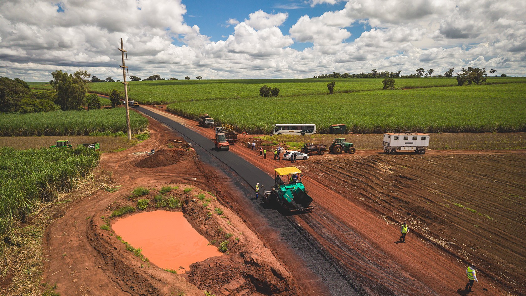 Perspectiva aérea de trabalhos de pavimentação de asfalto em meio a canaviais, com uma pavimentadora Vögele e um cilindro Hamm no centro, junto a outras máquinas de construção.