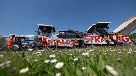 Material feeders and pavers paving asphalt behind a meadow full of daisies.