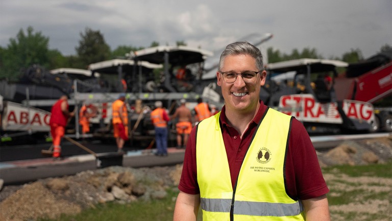 Mario Peiker, Managing Director and Technical Director of Hart Consult International GmbH, smiles at the camera as asphalt paving goes on behind him.