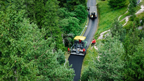 Una finitrice esegue la posa in opera di uno strato di base su una stretta strada di montagna. Superfici boschive su entrambi i lati della strada.