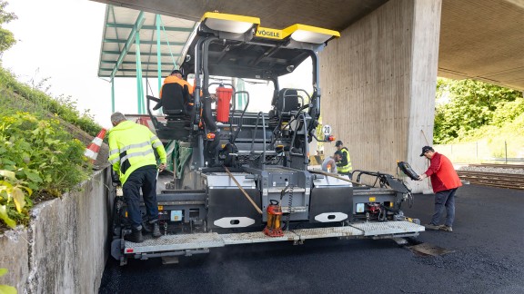 Two large light modules shine from the rear of the paver roof onto the screed and the paved asphalt.