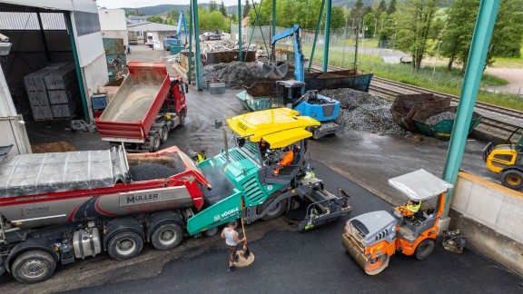 Paver being filled with mix; in the background, daily work at the recycling centre.