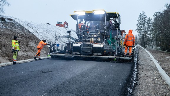 In light snowfall and twilight, the paving team pave the country road. The paver’s LED lighting shines on the screed and the freshly paved road.