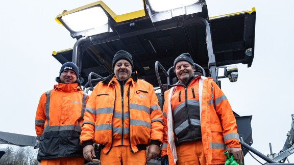 Three gentlemen smile contentedly into the camera. The cabin of the Vögele paver is visible behind them.