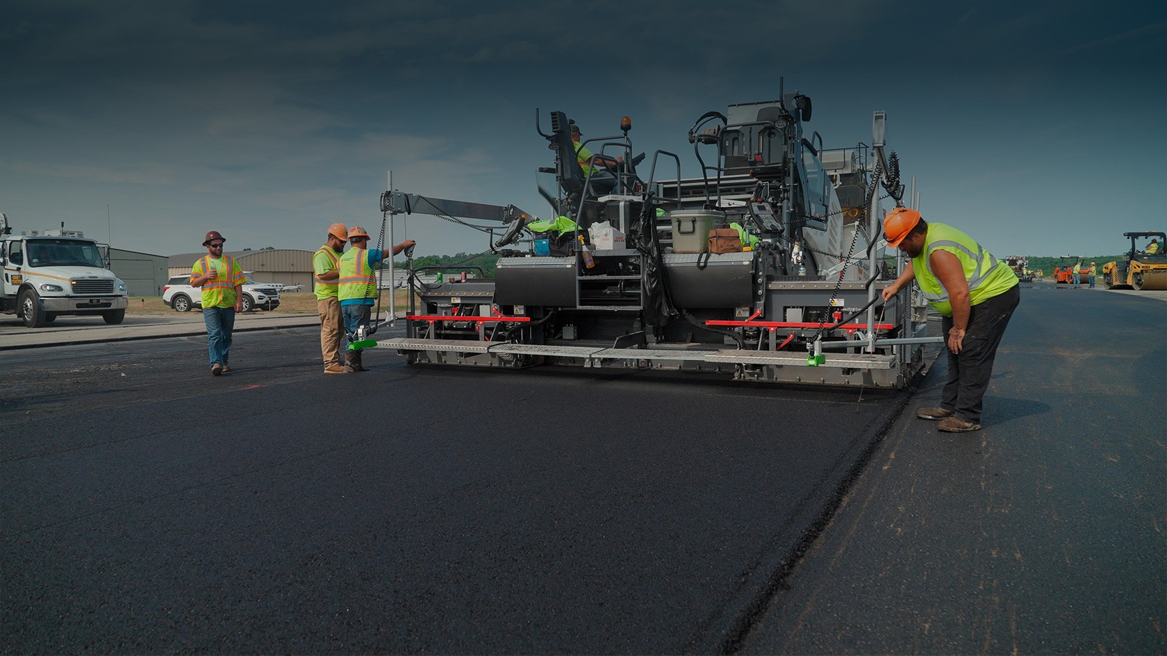 Le finisseur Vögele SUPER 2000-3i lors de la pose de la couche portante en enrobé Au premier plan, la nouvelle couche portante, à l’arrière-plan, le finisseur. Des travailleurs contrôlent le résultat.