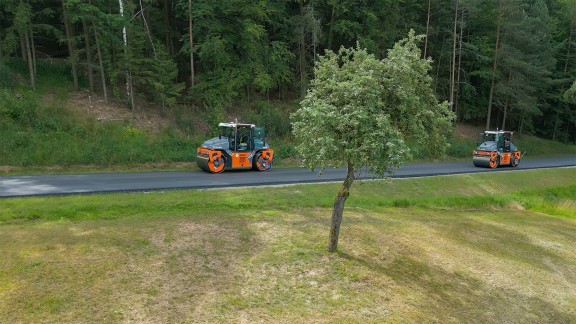 Two tandem rollers compacting a freshly asphalted country road, woodland coming into view in the background.