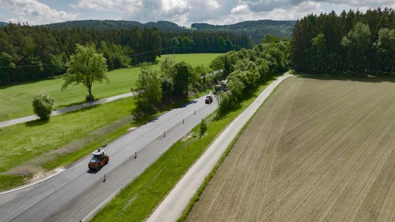 A bird’s eye view of the production system, comprising a paver and two rollers asphalting and compacting a country road at the same time.