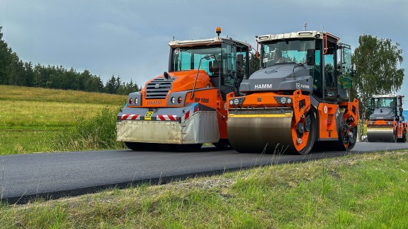 Two tandem rollers compacting a freshly asphalted country road, the edge of woodland in the background.