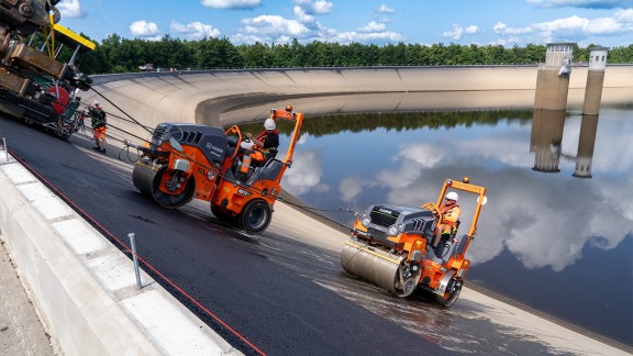 Two rollers compact the freshly laid asphalt on a steep surface.