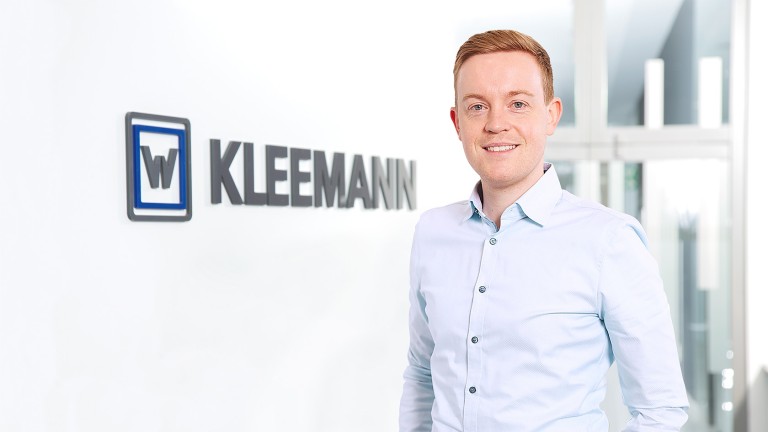 Laurin Winter, Product Manager di frantoi a urto presso Kleemann: Un giovane biondo in abito da ufficio si trova accanto al logo Kleemann e sorride alla telecamera.