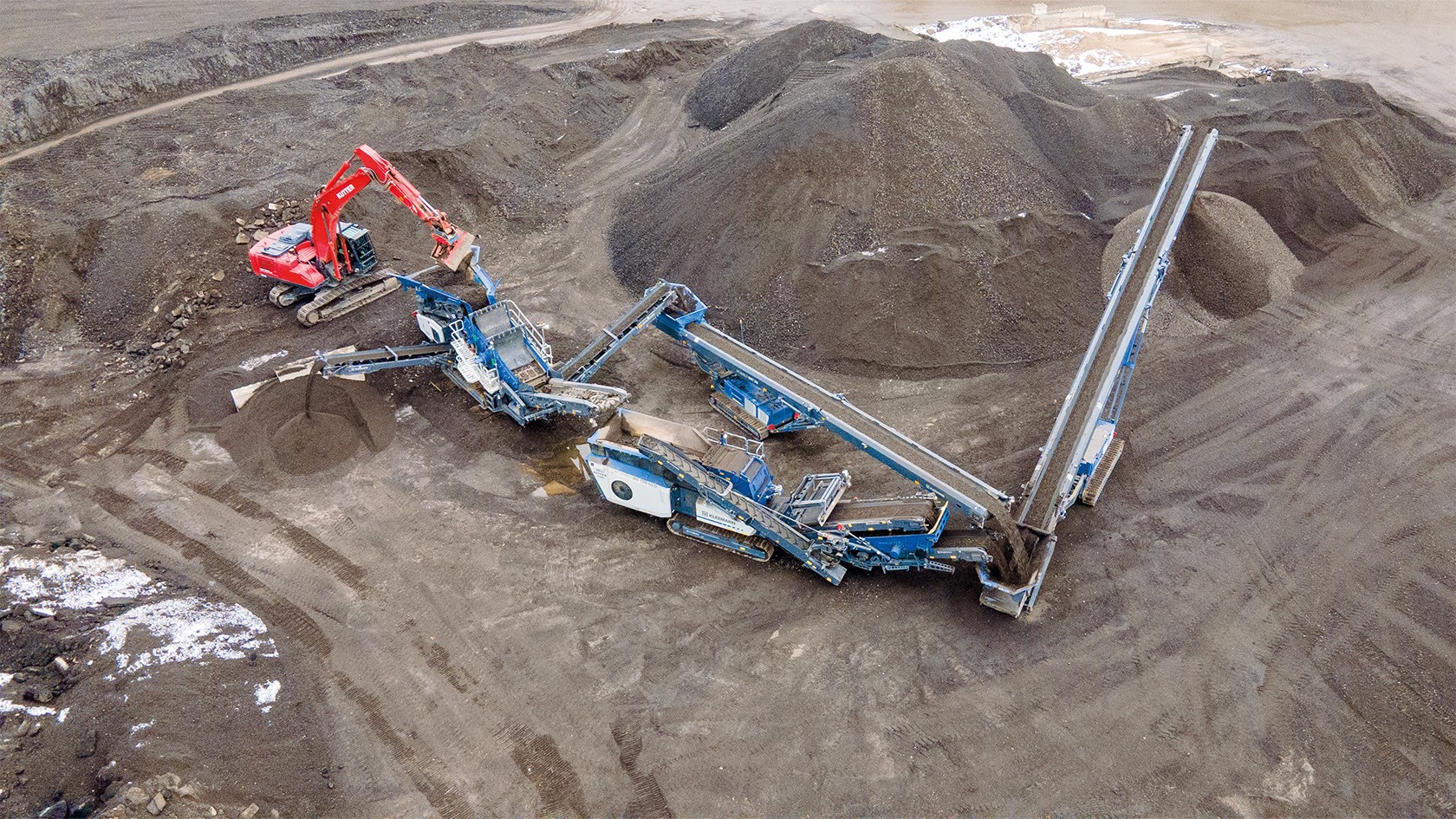 La cadena de instalaciones de Kleemann se encuentra en un centro de reciclaje de asfalto viejo y es alimentada por una excavadora roja.