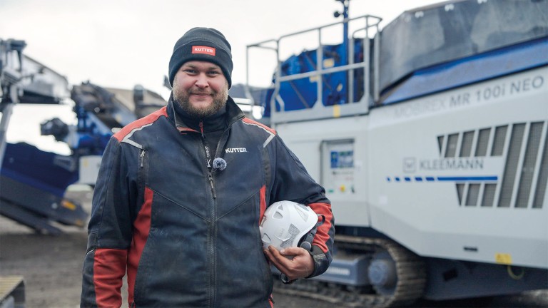 Retrato de un hombre con barba frondosa, de pie delante de una trituradora de impacto, sonriendo a la cámara. Bajo el brazo llevaba su casco de seguridad.