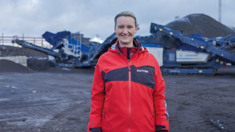 Mujer rubia - Simone Eichiner, directora de instalaciones mezcladoras de asfalto en Kutter - Con una chaqueta roja brillante, sonríe directamente a la cámara.