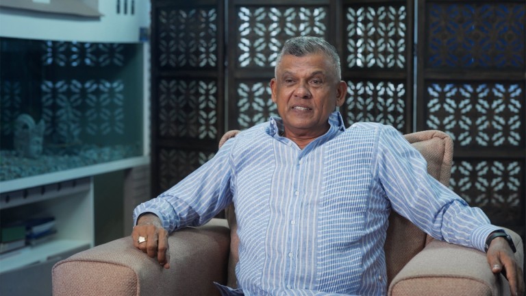 Brian Tiwarie, a middle-aged gentleman, sits in a light-coloured armchair and smiles at the camera. Behind him a mobile partition wall with carvings.