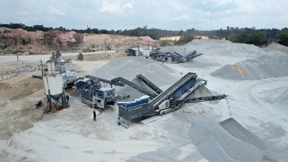 Aerial image of quarry in which grey natural stone is being processed. You can see a plant train from Kleemann and a light-brown quarry edge behind it. The beginning of the jungle at the very back.
