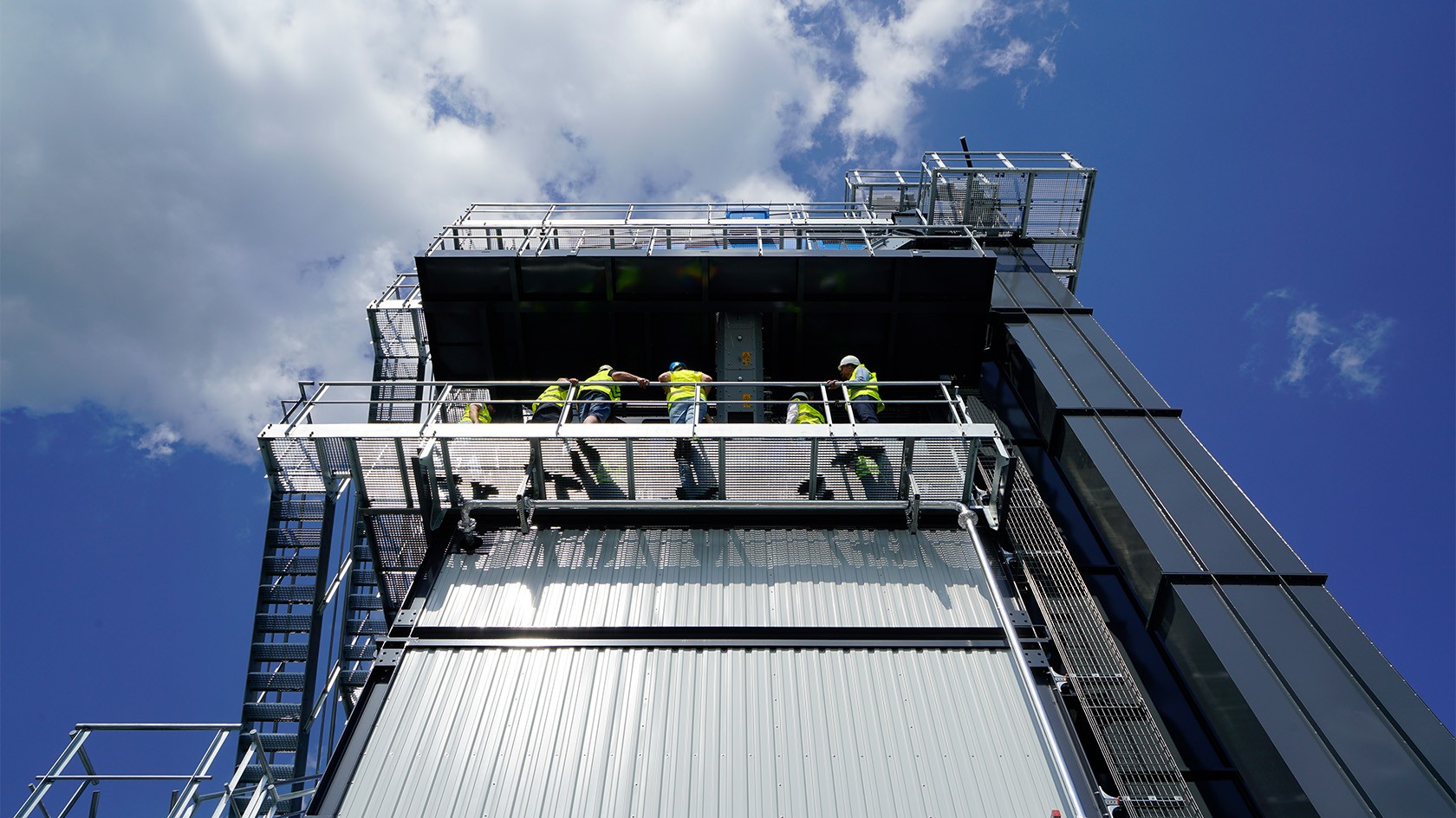 Asphalt mixing plant viewed from below at an angle. Five trainees are seen standing high up at the railings.