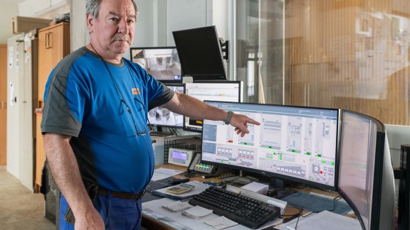 Mixing foreman standing in front of a desk, pointing at the screen of the computer that is on the desk.