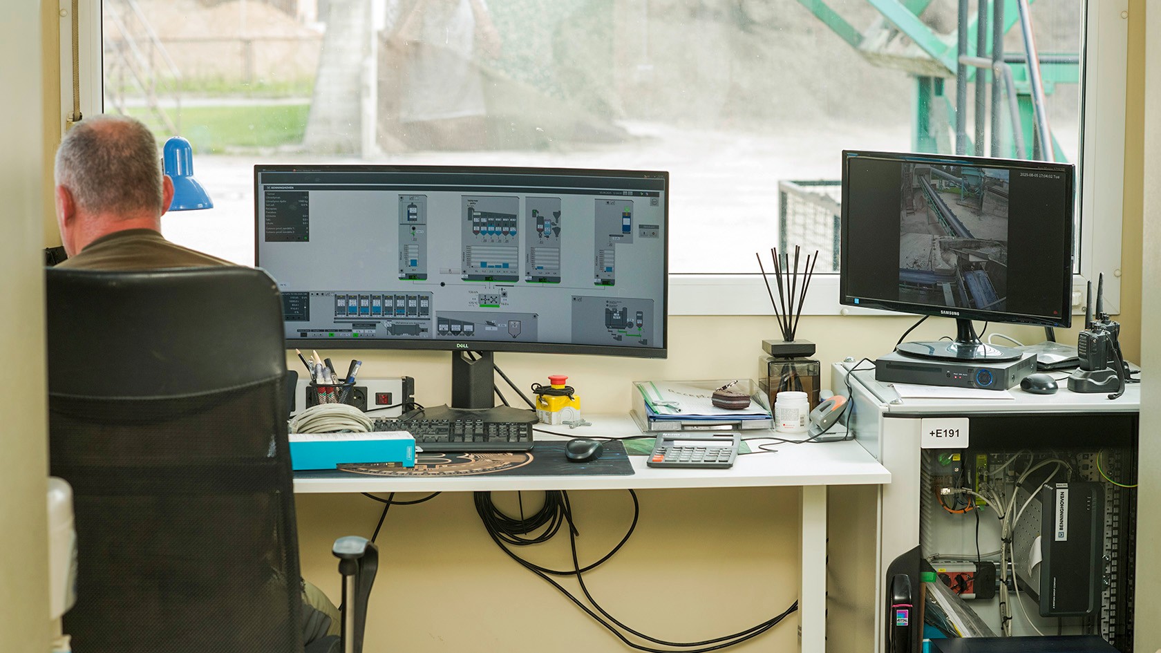 Mann sitzt am Schreibtisch. Auf dem Computerbildschirm wird die Steuerung der Asphaltmischanlage angezeigt.